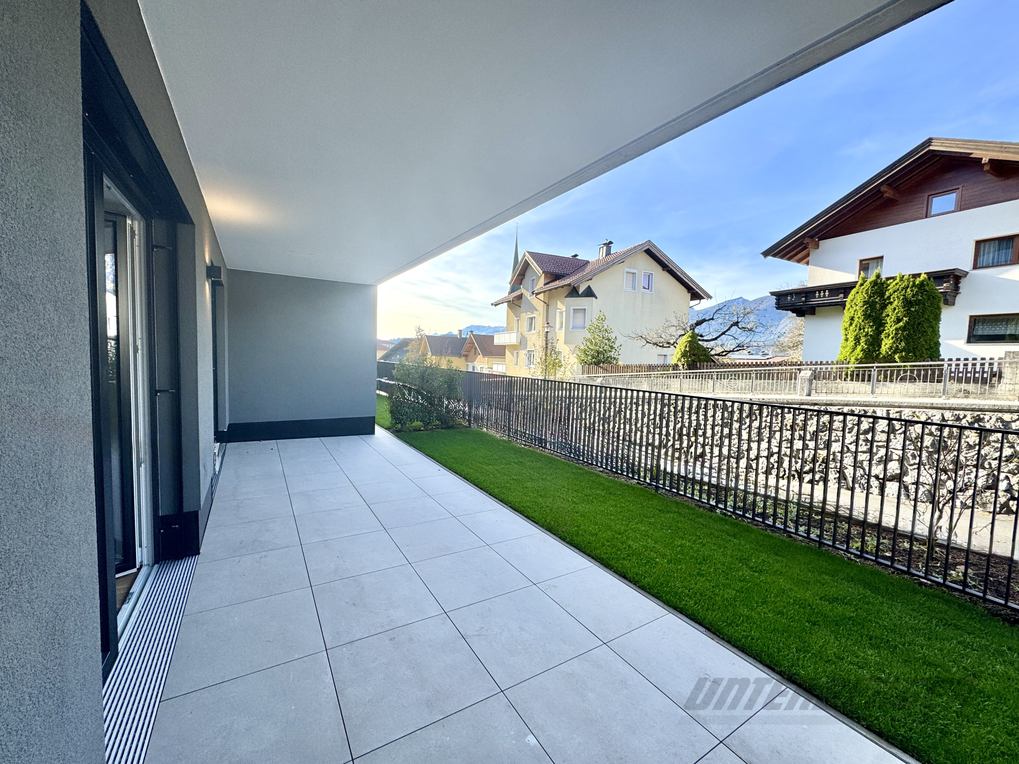 Großzügige, überdachte Terrasse mit grauen Fliesen, Rasenstreifen und Blick auf die Berge