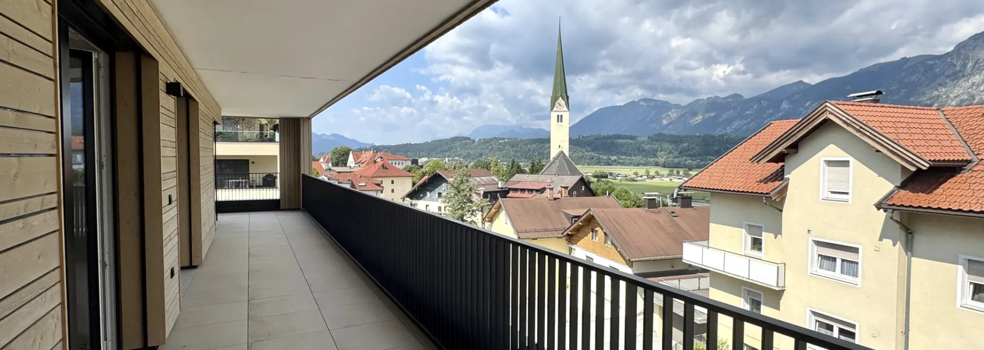 Balkon mit Holzfassade, grauen Fliesen und Süd-West-Blick auf Kirchturm und Bergpanorama